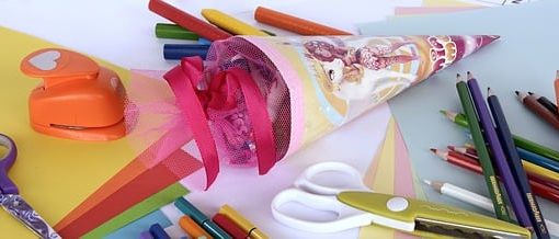 Eine selbstgebastelte Schultüte liegt zwischen stiften scheren und Buntem Papier auf einem tisch